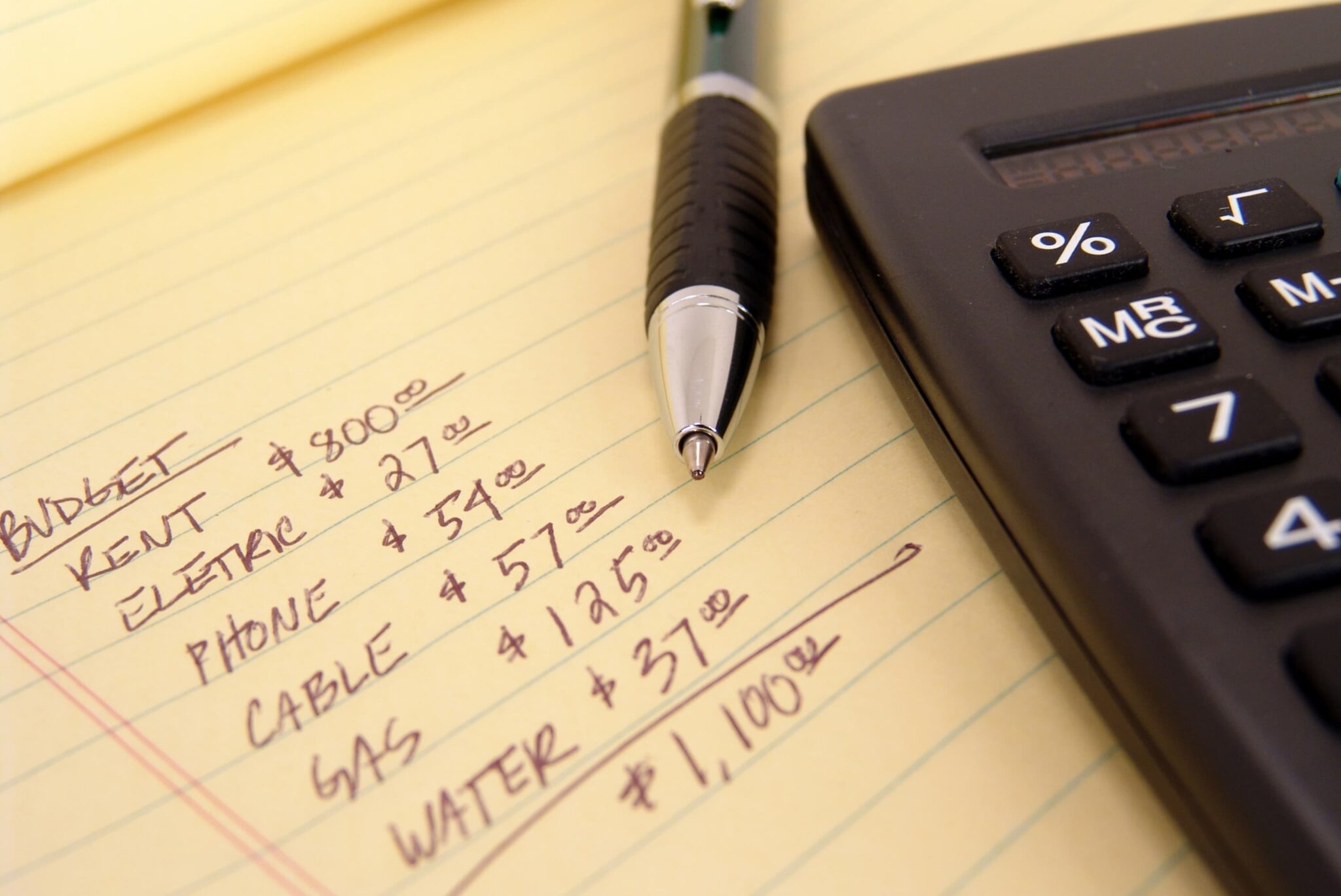 A pen rests on a handwritten budget list on a yellow paper, showing expenses for rent, utilities, and totaling $1,100. A black calculator is partially visible on the right.