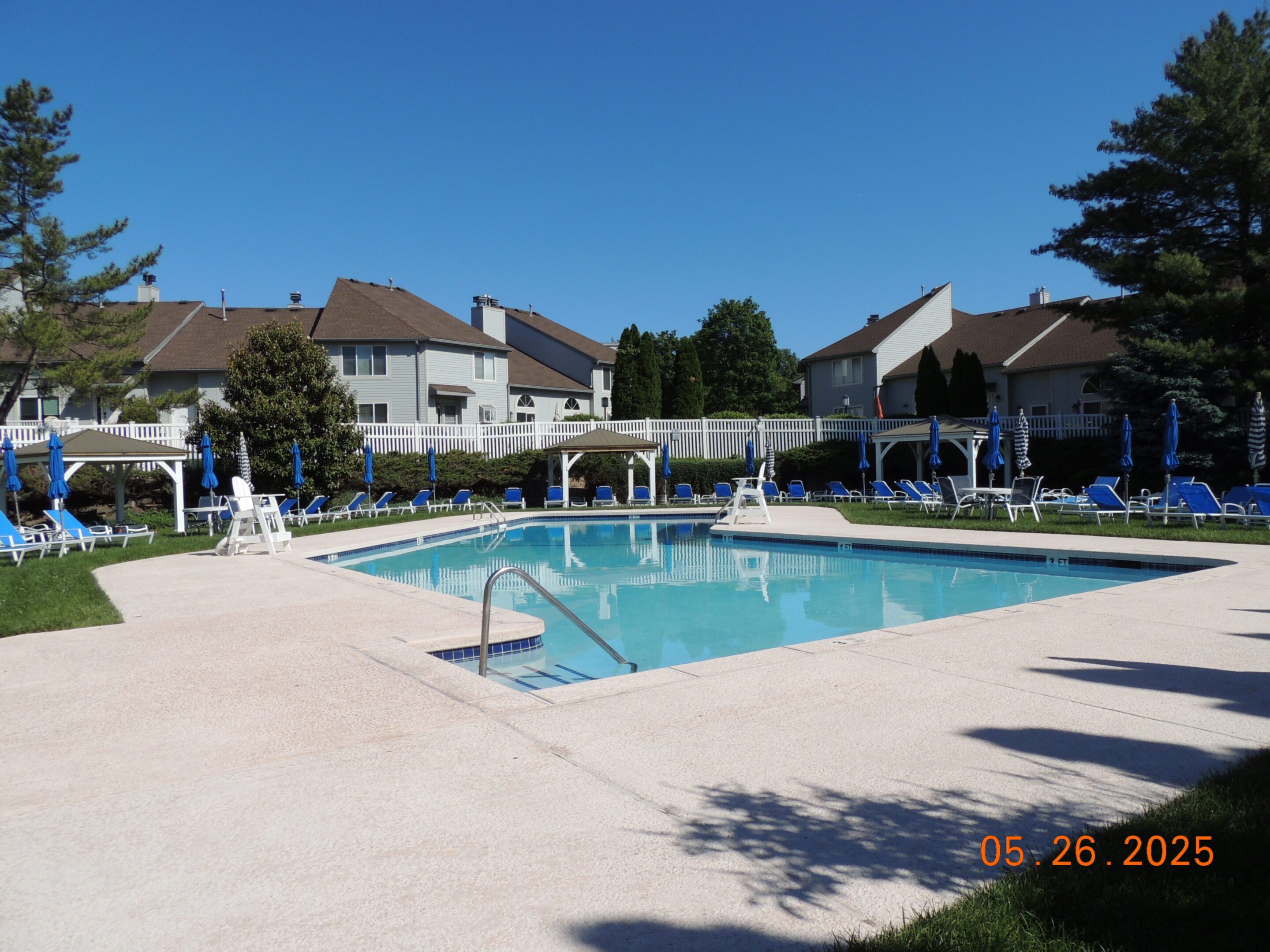 Outdoor swimming pool surrounded by lounge chairs and umbrellas in a residential community on a sunny day. Date stamp on image reads May 26, 2025.