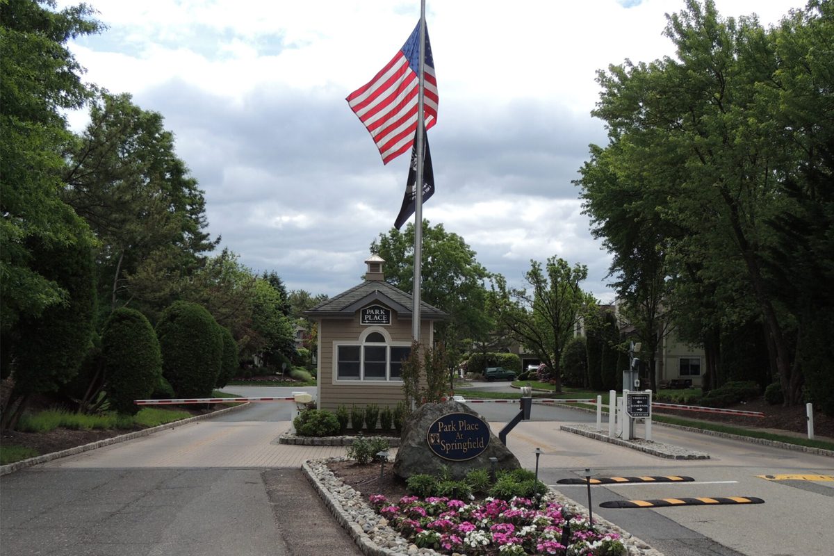 re-img A gated entrance to Park Place at Springfield features a security booth, flower bed, and flagpole with American and POW/MIA flags. Trees and houses are visible in the background.